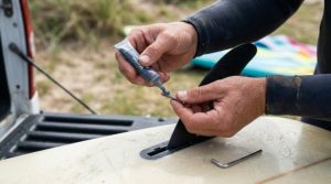 Close-up of a surfer applying anti-seize lubricant to a surfboard fin screw before inserting it into the fin box